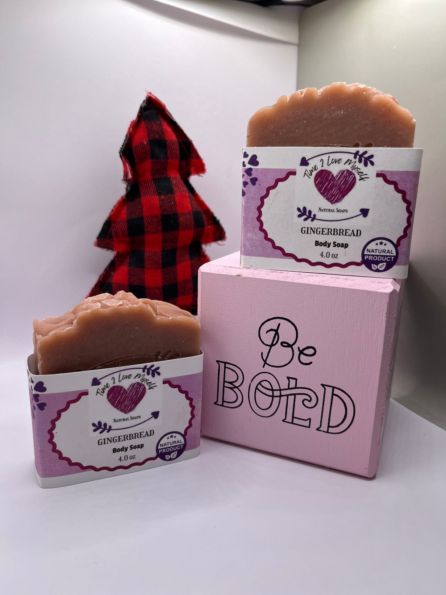 Two bars of handmade gingerbread soap, placed on a surface with a pine in the background and a square box saying Be Bold.